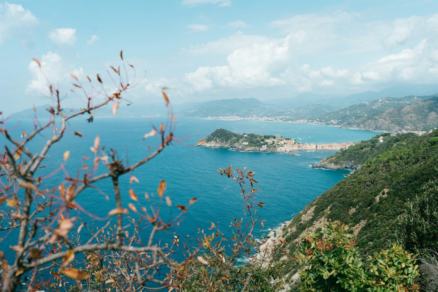 Costa ligure vista dall'alto tra rami dai colori d'autunno, con il mare azzurro e un promontorio con un borgo